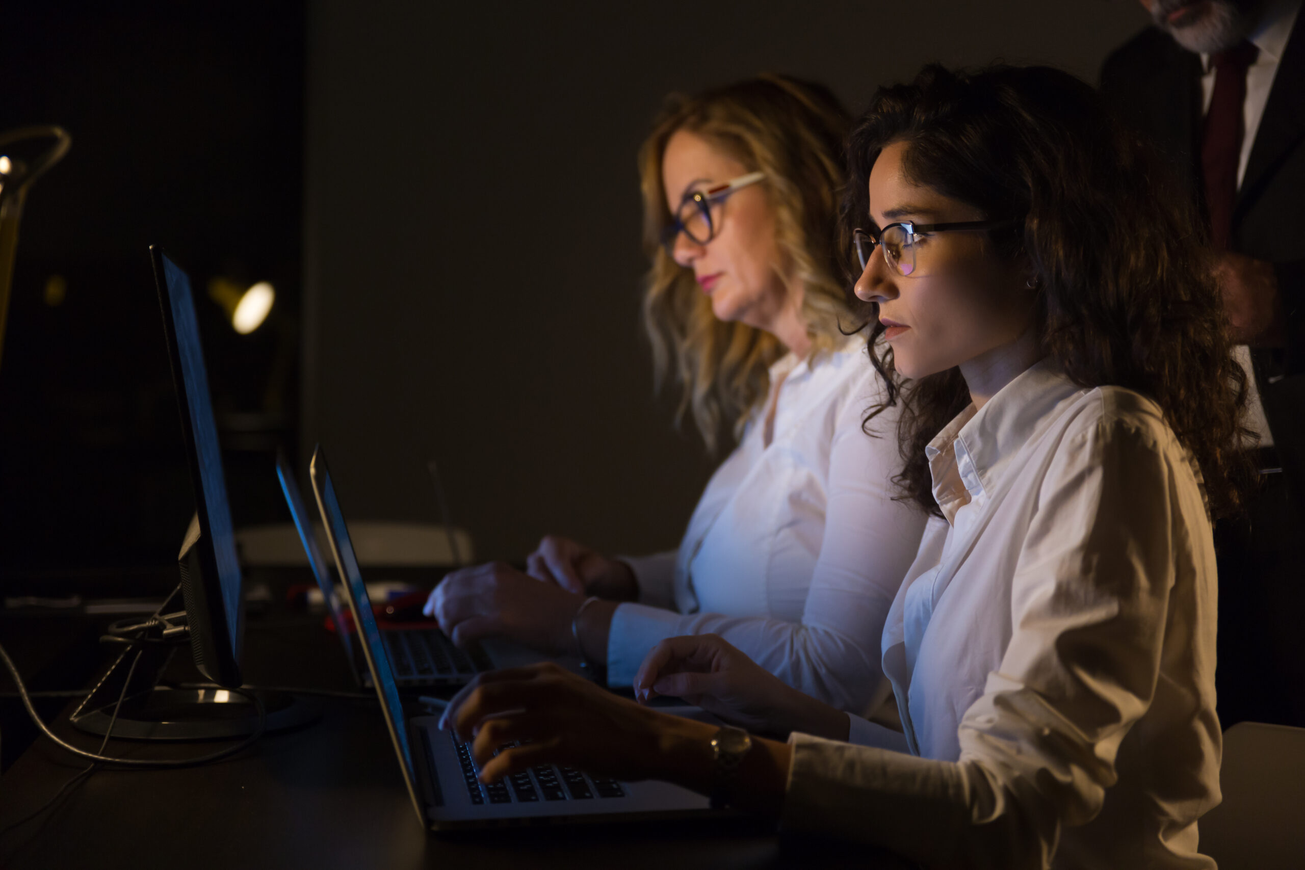 Serious businesswomen using laptops. Side view of two focused young businesswomen working with laptop computers in dark office. Working late concept